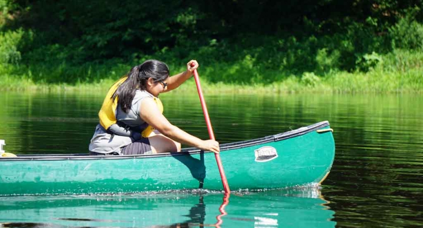 A person wearing a life jacket paddles a green canoe. There is a green shoreline in the background. 